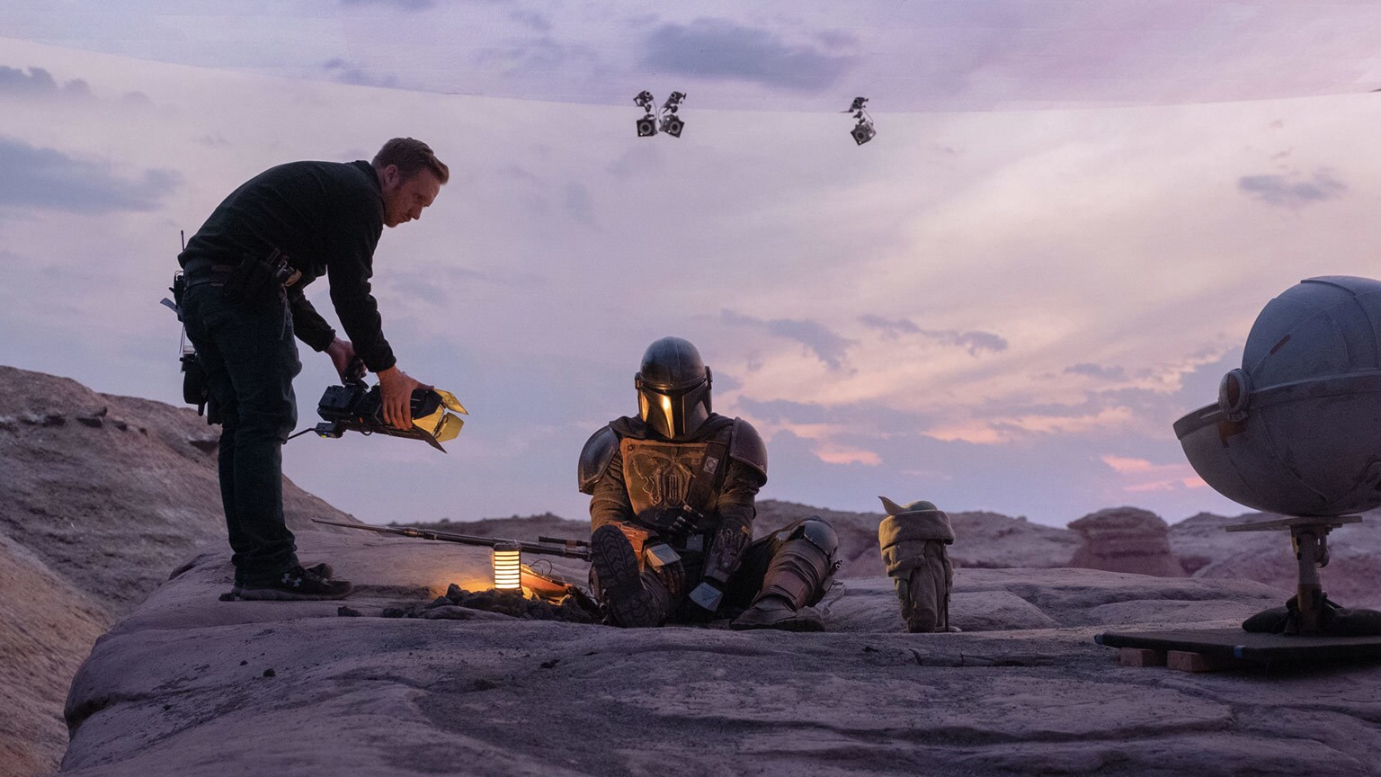 Filmmaker crouches with a camera on a rocky desert set, filming a seated armored Mandalorian at dusk.