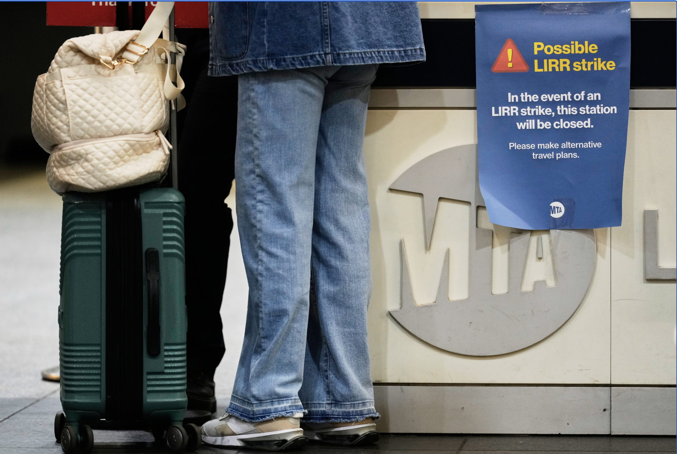 Two travelers with a green rolling suitcase stand at a counter beside a blue sign warning of a possible LIRR strike.
