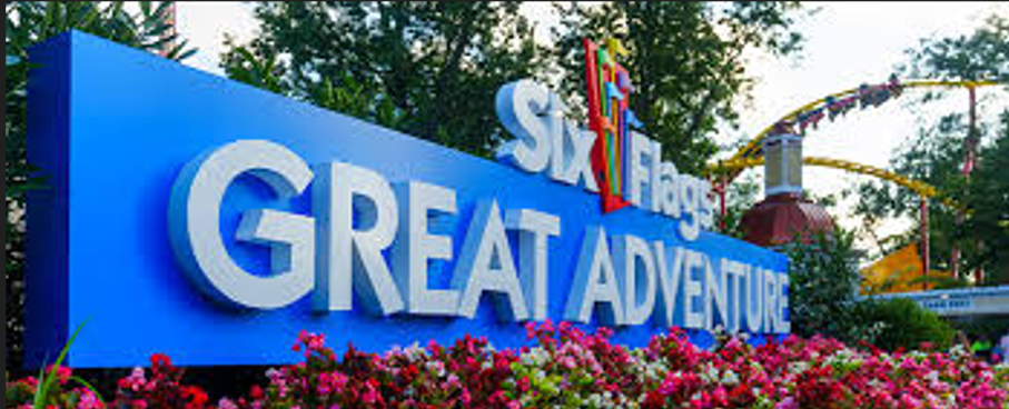 Six Flags Great Adventure sign with a blue background, surrounded by pink flowers and a distant roller coaster.