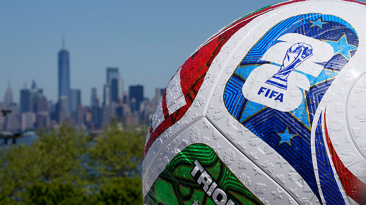Close-up of a FIFA World Cup soccer ball with a colorful design, set against the New York City skyline in the distance, including tall buildings and a blue sky.