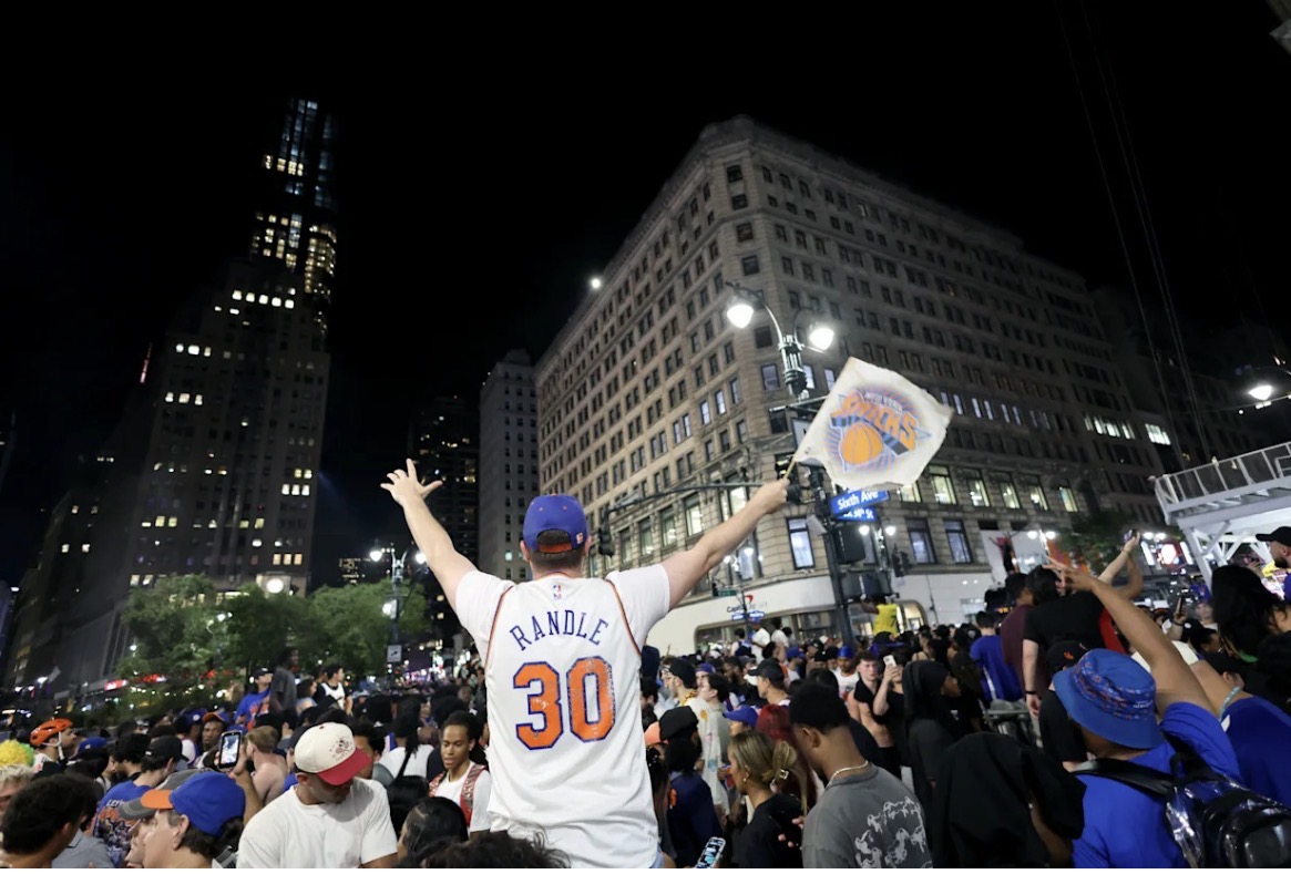 Crowd celebrating at night on a city street; man in a white jersey with 'RANDLE 30' on the back raises his arms while waving a flag with a basketball logo.