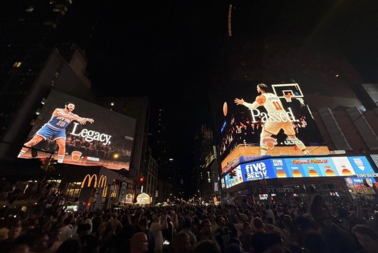 Nighttime Times Square scene with enormous basketball-themed billboards reading 'Legacy' and 'Passed,' and a crowded street below.