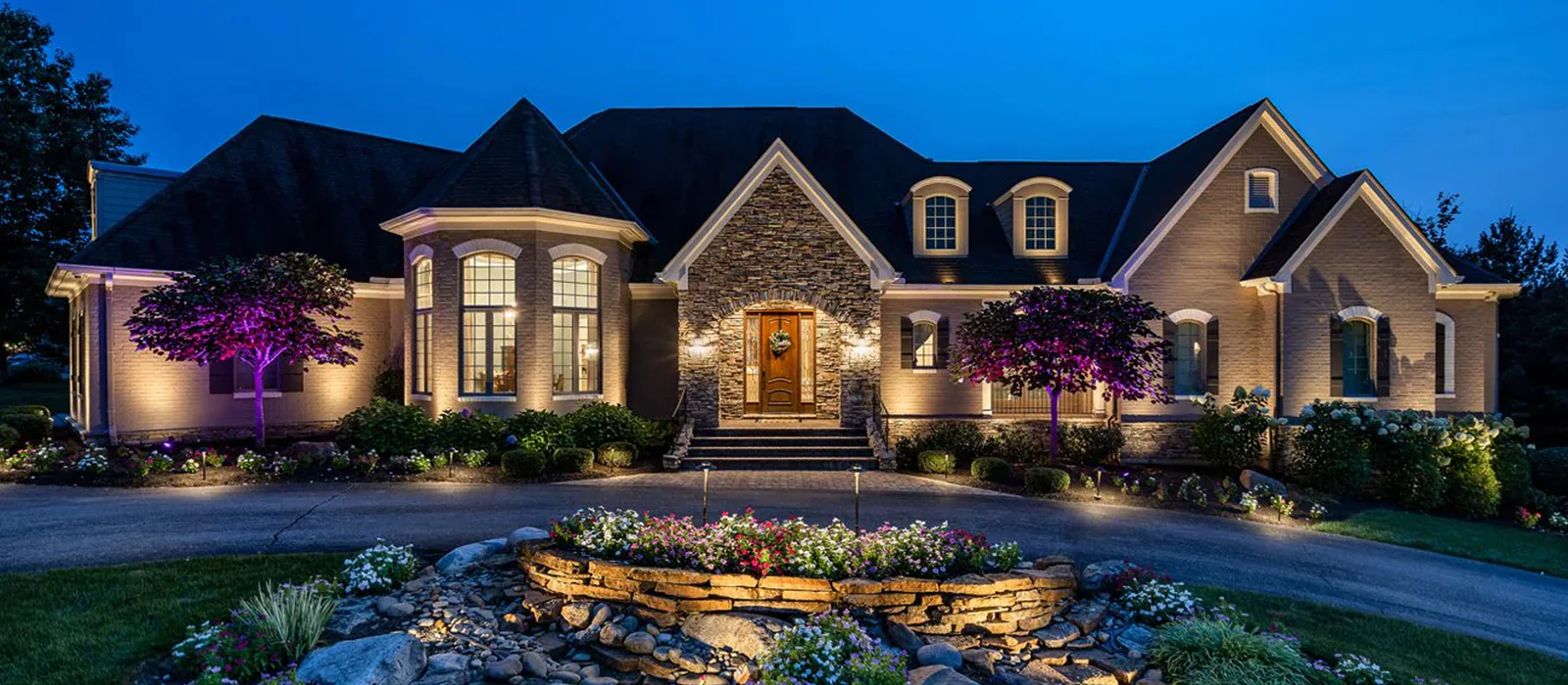 Evening view of a large, well-lit house with stone facade and landscaped front yard, including a circular stone flower bed in the foreground