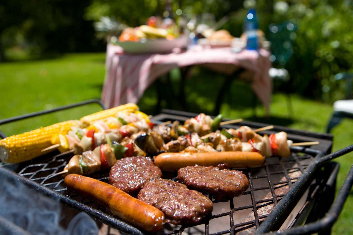 Barbecue grill with burgers, sausages, corn on the cob and veggie skewers cooking outdoors; blurred picnic table in background.