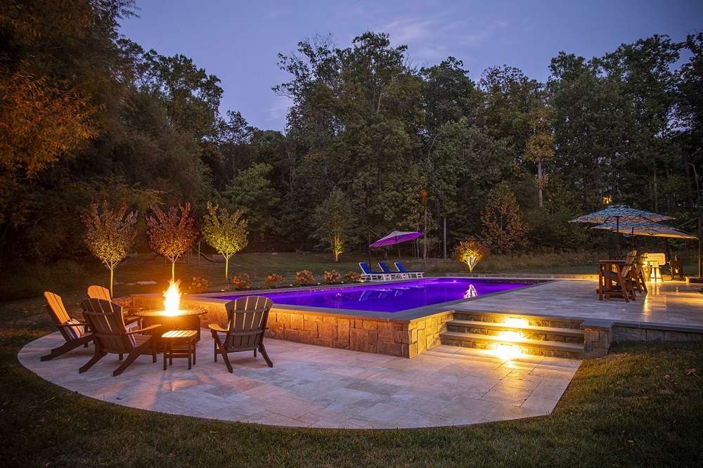 Backyard pool area at dusk with a lit fire pit, wooden chairs, and a purple-lit pool surrounded by trees.