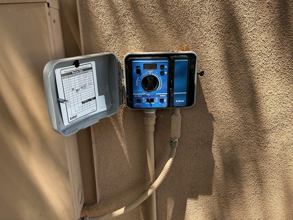 Blue irrigation timer with a digital display and large control dial, mounted on a textured beige wall with the weatherproof cover open and a label inside the door.