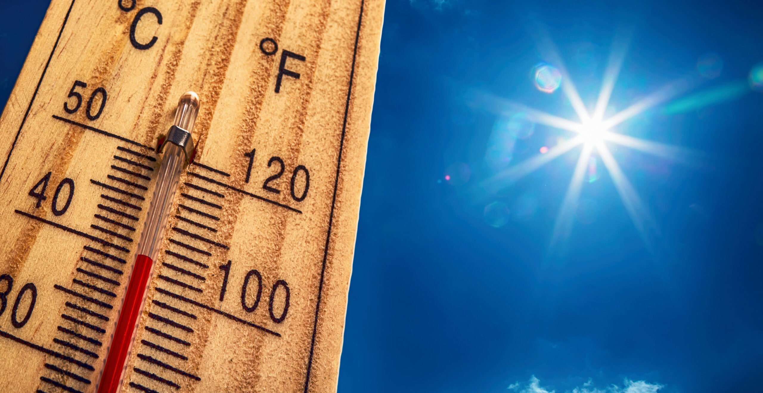 Close-up of a glass thermometer on a wooden ruler with a bright sun in a clear blue sky.