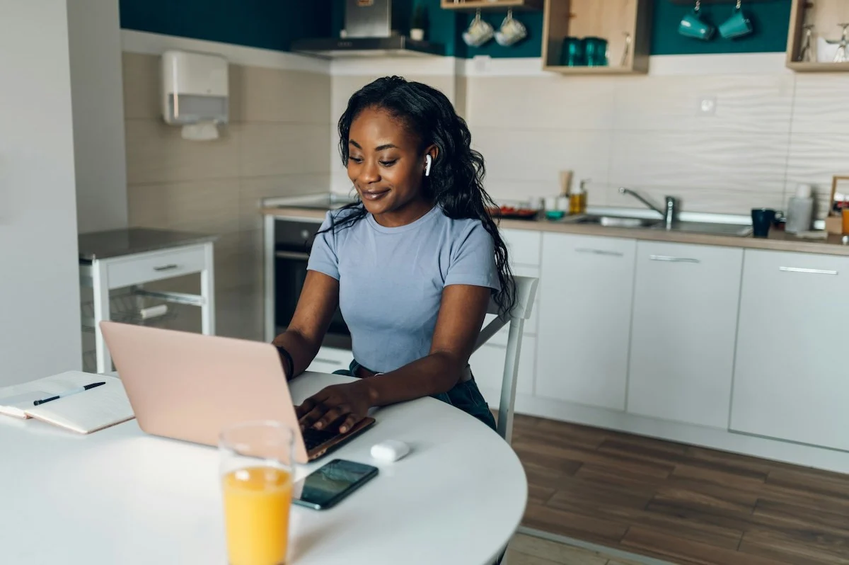 Woman in a blue shirt typing on a pink laptop at a white round table in a modern kitchen, with a glass of orange juice nearby.